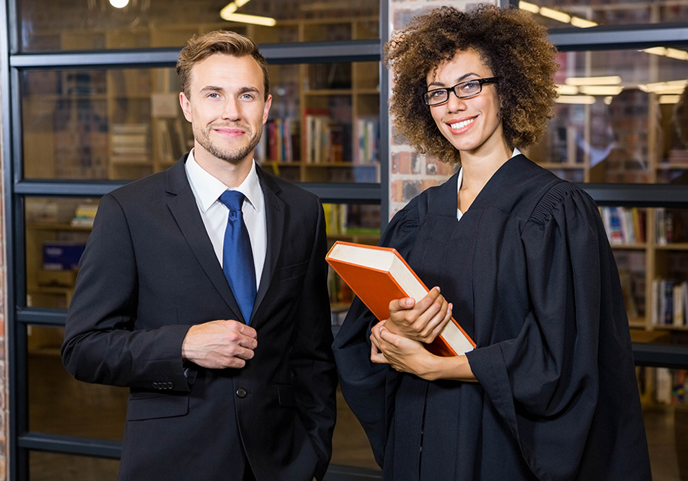Two people in a library, one in a suit and the other in an academic gown, holding a book.