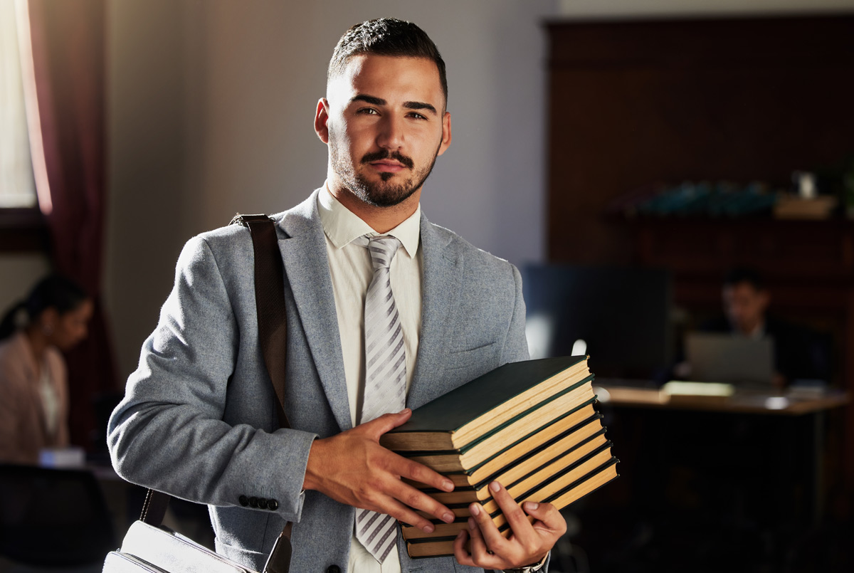 Person in a gray suit holding a stack of books in a warmly lit room with others working in the background.